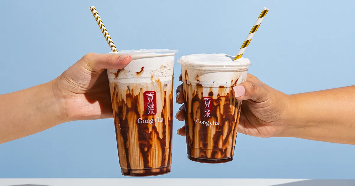 Two hands clinking Gong Cha chocolate-drizzle milk teas with frothy tops and striped paper straws against a pale blue background.