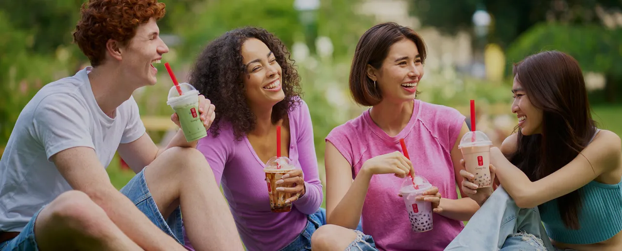 Four friends sitting on grass, smiling and holding bubble tea drinks in a park. They appear relaxed and cheerful, enjoying a sunny day.