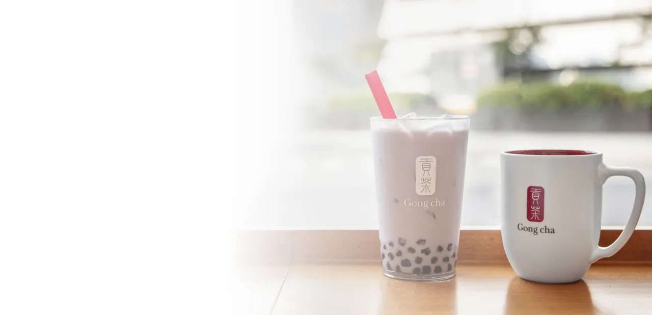 Bubble tea with tapioca pearls and a red straw, next to a white Gong Cha mug, placed on a wooden counter by a window.