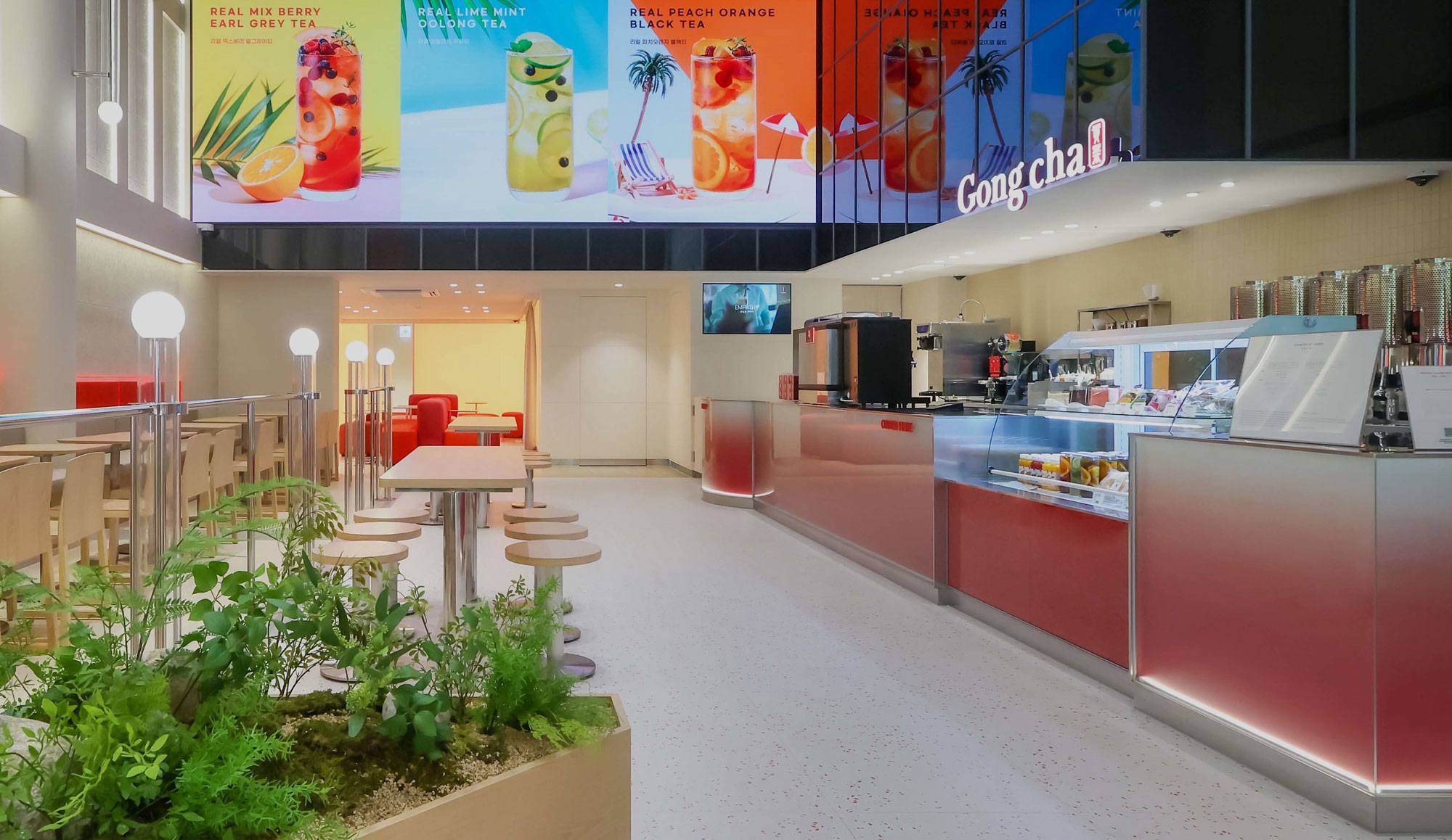 Bright Gong Cha interior: sleek service counter, refrigerated display, high stools, communal tables, planter, and colorful drink banners.