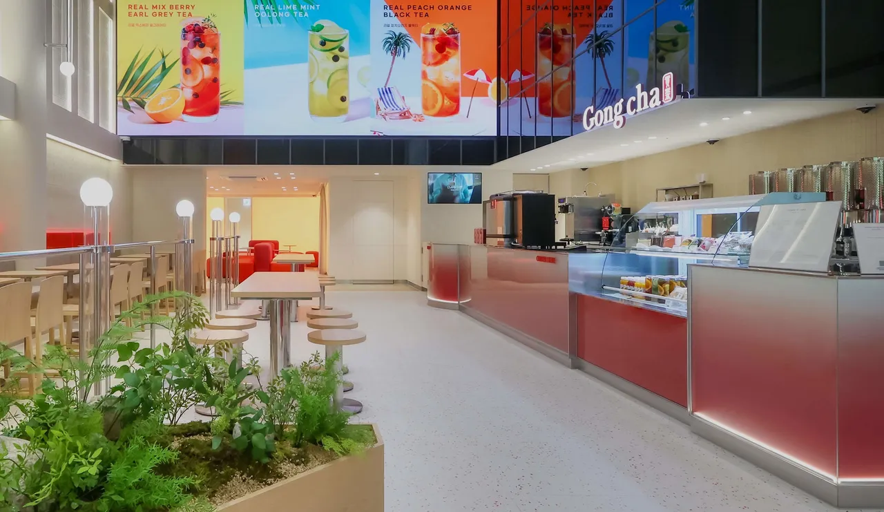 Bright Gong Cha interior: sleek service counter, refrigerated display, high stools, communal tables, planter, and colorful drink banners.