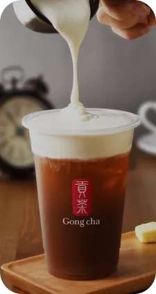 A hand pouring creamy milk into a cup of iced tea from Gong cha, with an alarm clock and sugar cubes in the background.