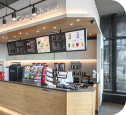 Modern bubble tea shop interior with a wooden counter, digital menu displays, and beverage preparation area. Bright and inviting atmosphere.
