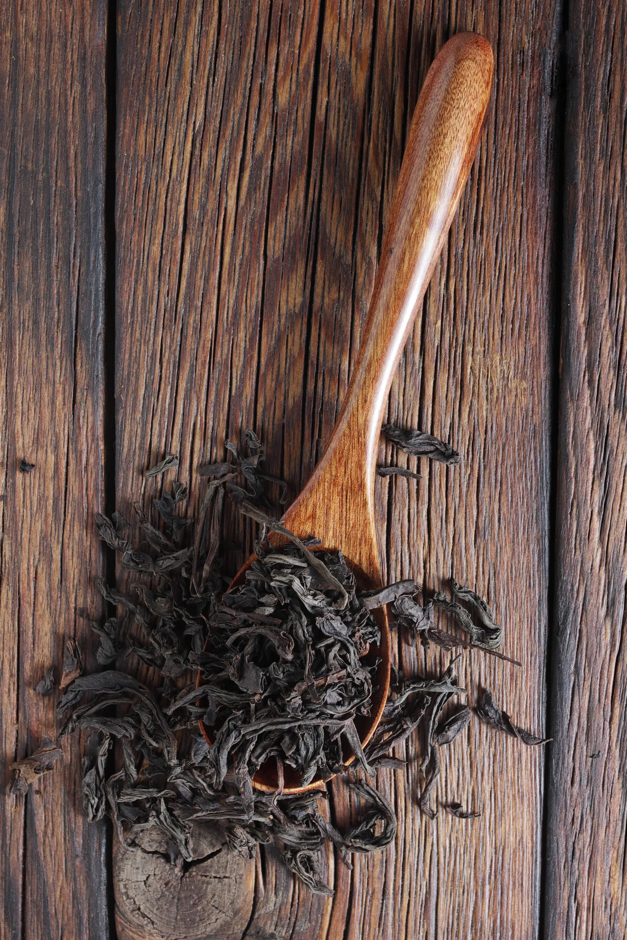 A wooden spoon filled with dried tea leaves rests on a rustic wooden surface.