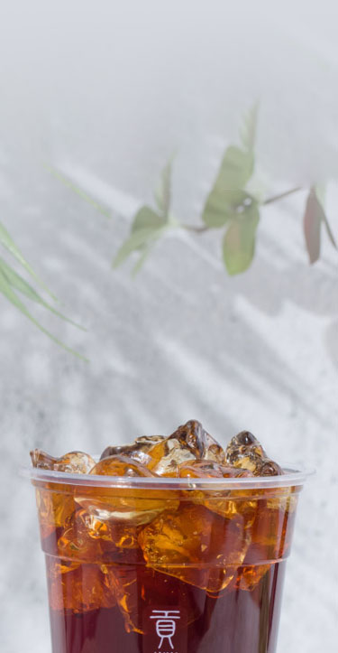 A cup of iced tea with a clear lid, filled with ice cubes. Blurred greenery and a soft background create a refreshing feel.