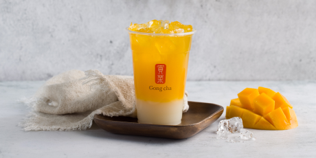 A layered mango bubble tea with ice in a clear cup on a wooden tray, accompanied by a towel, ice cubes, and fresh mango slices.