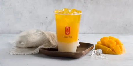 A layered mango bubble tea with ice in a clear cup on a wooden tray, accompanied by a towel, ice cubes, and fresh mango slices.
