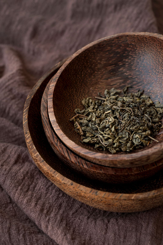 Dried tea leaves in a wooden bowl, stacked on two other bowls, placed on a textured brown fabric.