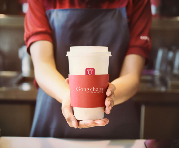 Person in a red shirt and apron holding a white cup with a red sleeve labeled "Gong cha" in a cafe setting.