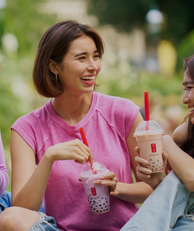Two people sitting outdoors, smiling and holding bubble tea with red straws. The person on the left wears a pink shirt.
