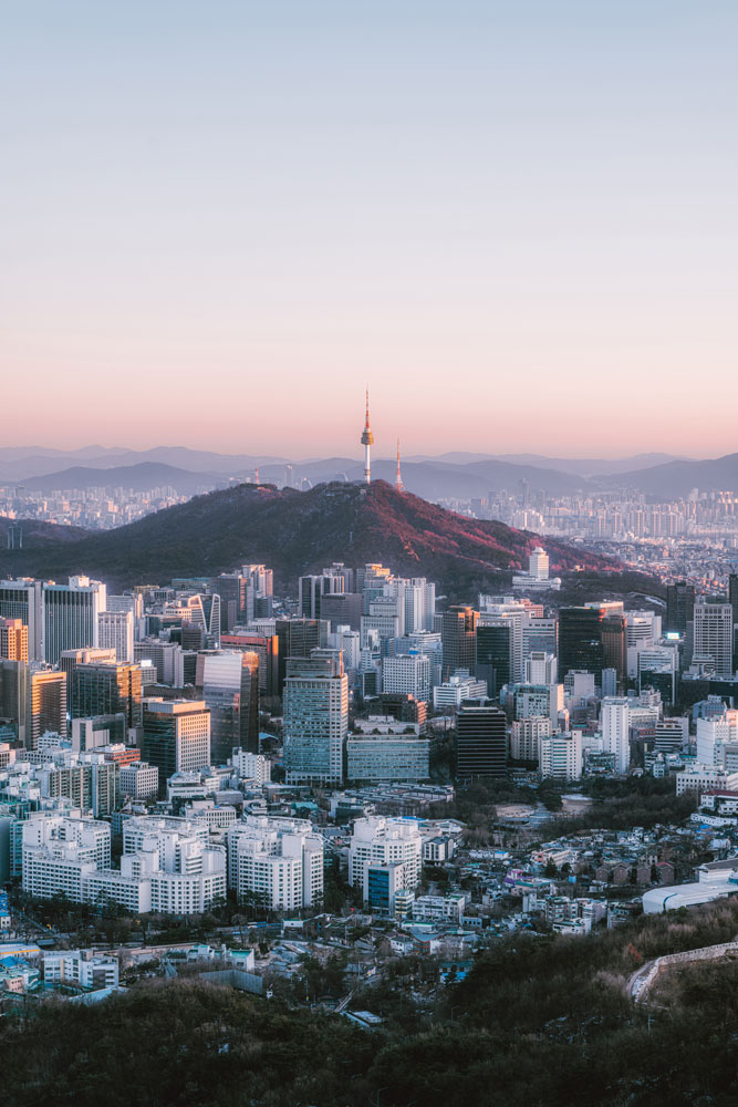 Aerial view of a cityscape at dusk, featuring a prominent tower on a hill surrounded by modern buildings and distant mountains.