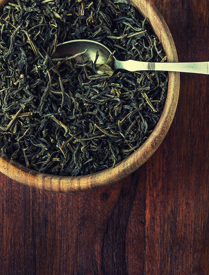 A wooden bowl filled with dried tea leaves and a metal spoon, placed on a dark wooden surface.