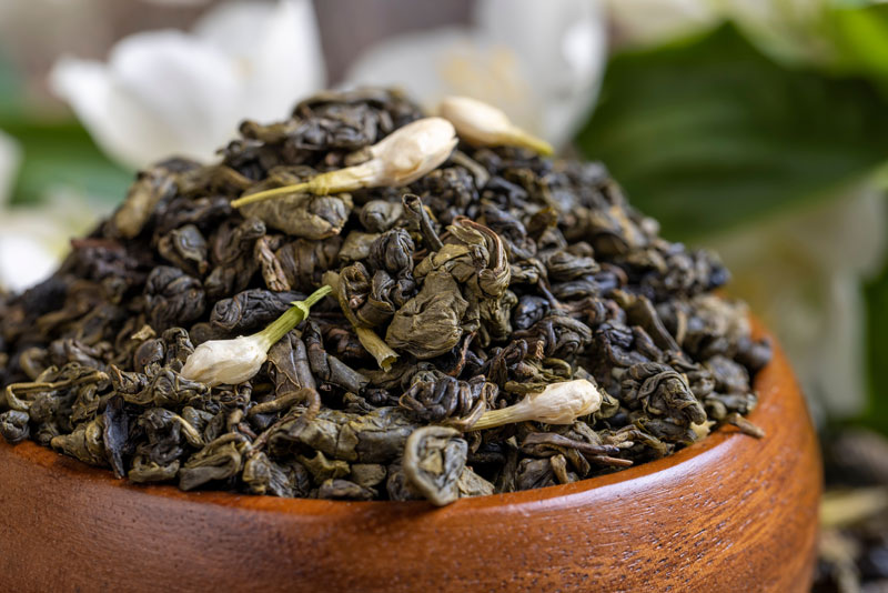 A wooden bowl filled with loose jasmine tea leaves and small jasmine flowers, with blurred white flowers in the background.