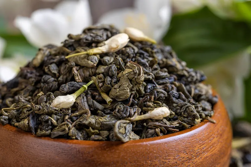 A wooden bowl filled with loose jasmine tea leaves and small jasmine flowers, with blurred white flowers in the background.