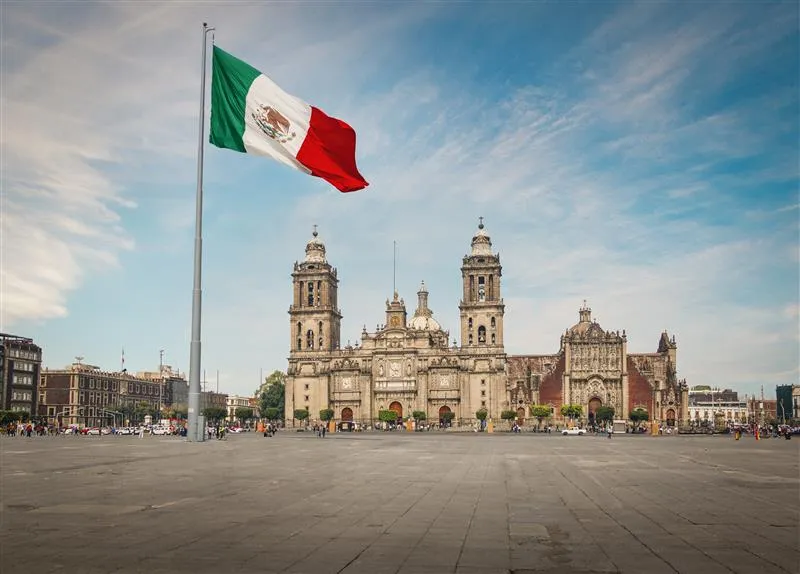 Mexican flag waving in front of the Metropolitan Cathedral at Zócalo, Mexico City, under a blue sky with scattered clouds.