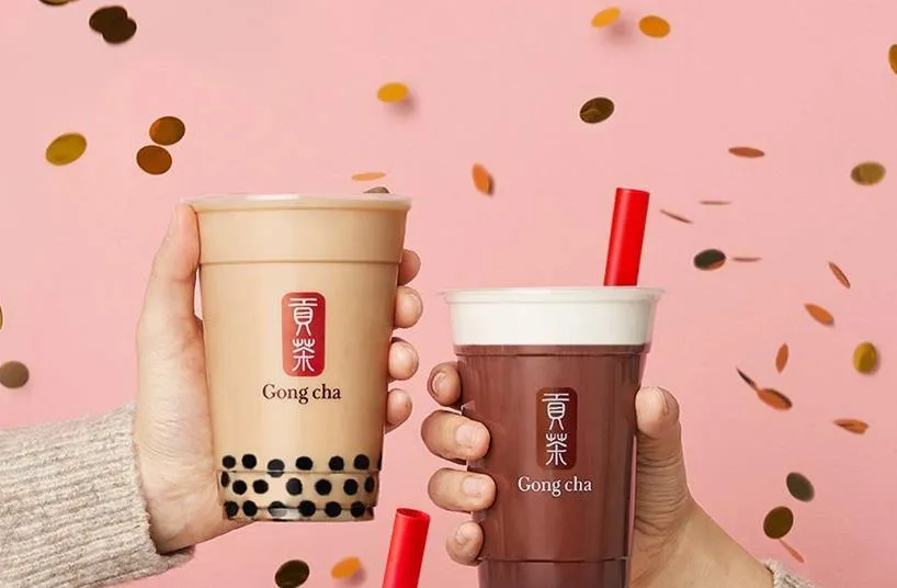 Two hands holding Gong Cha bubble tea cups with a pink backdrop and falling confetti.