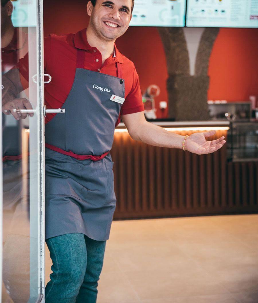 Smiling man in a red shirt and gray apron welcomes customers at a cafe entrance, with a modern interior visible in the background.
