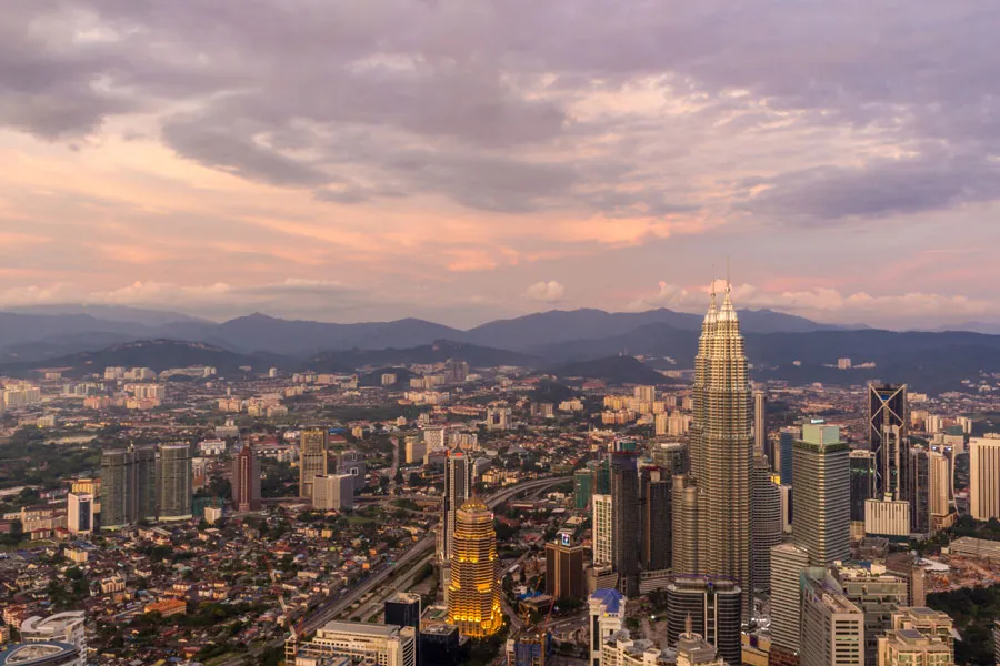 Aerial view of a cityscape at sunset, featuring tall skyscrapers, including a prominent twin-tower building, under a cloudy sky.