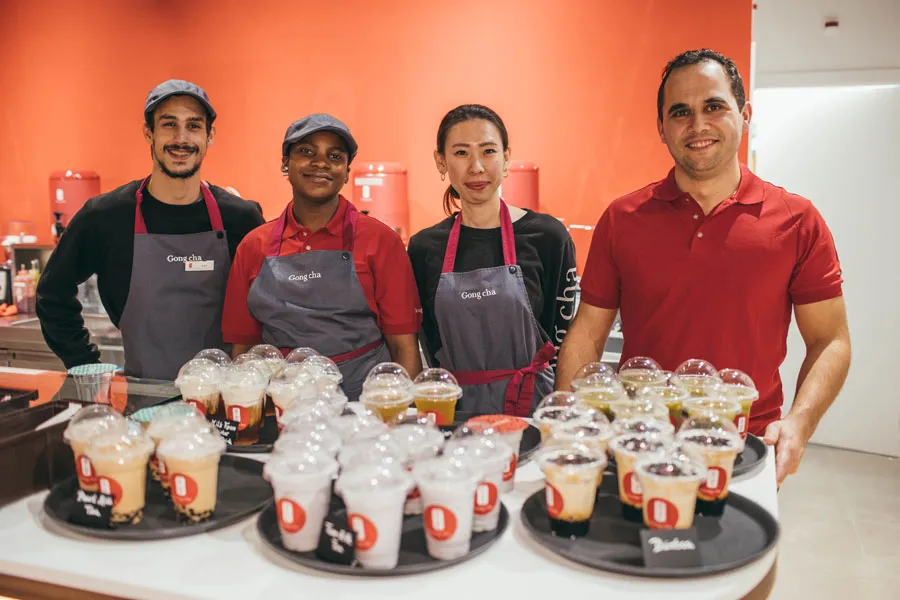 Four staff members in a cafe, wearing aprons, stand smiling with trays of drinks in front of a red wall.
