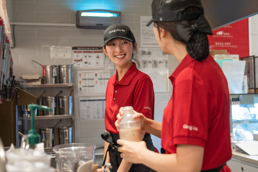 Two employees in red uniforms and black caps smiling and preparing drinks behind a counter in a cafe setting.