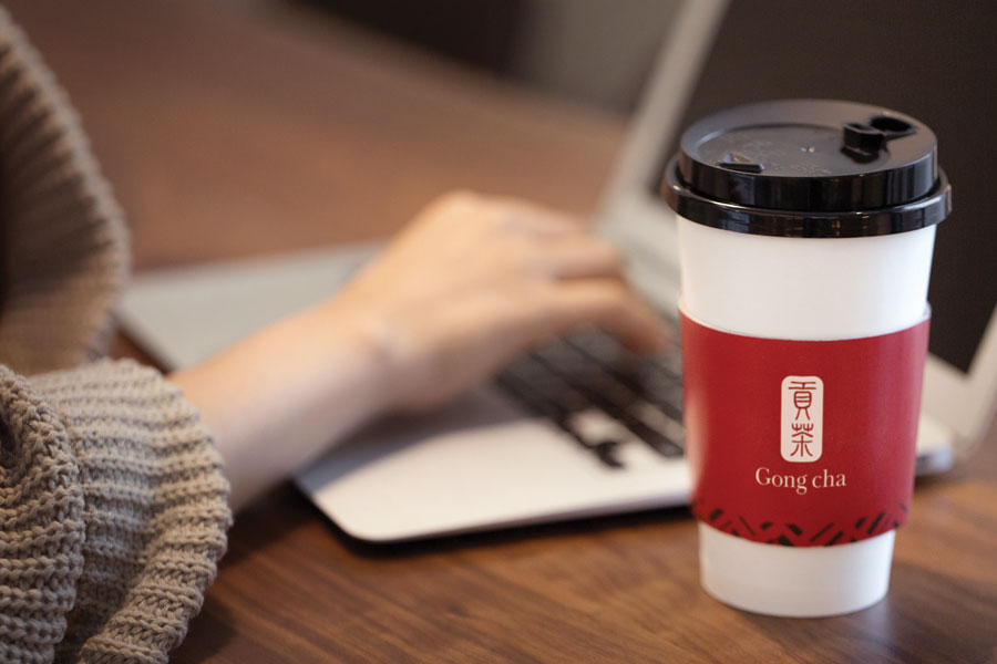 A person types on a laptop beside a Gong Cha cup with a red sleeve on a wooden table.