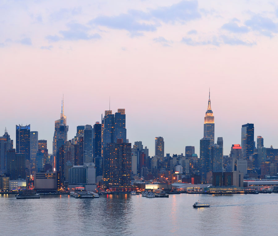 Skyline of a city at dusk with illuminated skyscrapers, including a prominent tower, reflected in a calm body of water.
