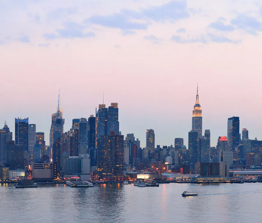 Skyline of a city at dusk with illuminated skyscrapers, including a prominent tower, reflected in a calm body of water.