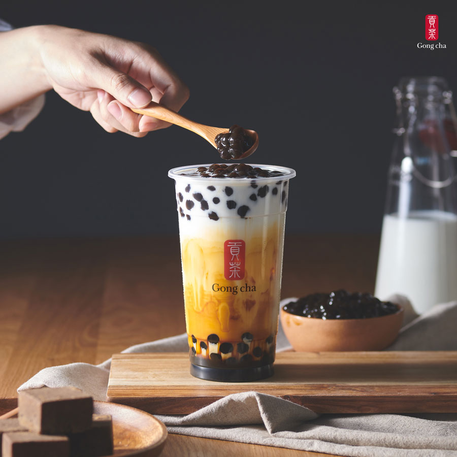 A hand holds a spoon with tapioca pearls over a bubble tea cup, labeled "Gong cha," on a wooden board with milk and sugar cubes nearby.
