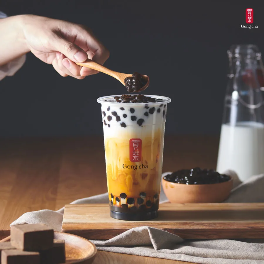 A hand holds a spoon with tapioca pearls over a bubble tea cup, labeled "Gong cha," on a wooden board with milk and sugar cubes nearby.