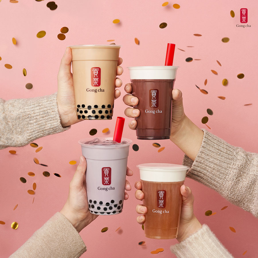 Four hands holding different bubble tea cups with various toppings, set against a pink background with floating confetti.