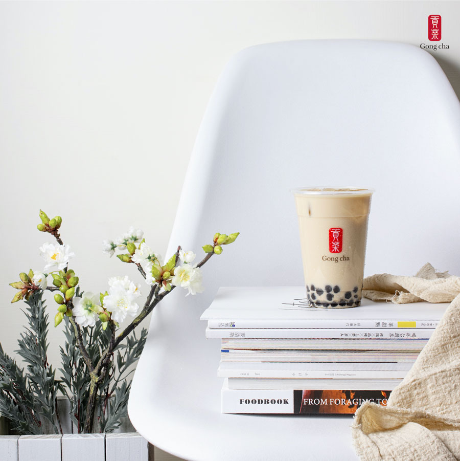 A cup of bubble tea on stacked books beside a flowering branch, all placed on a white chair against a plain background.