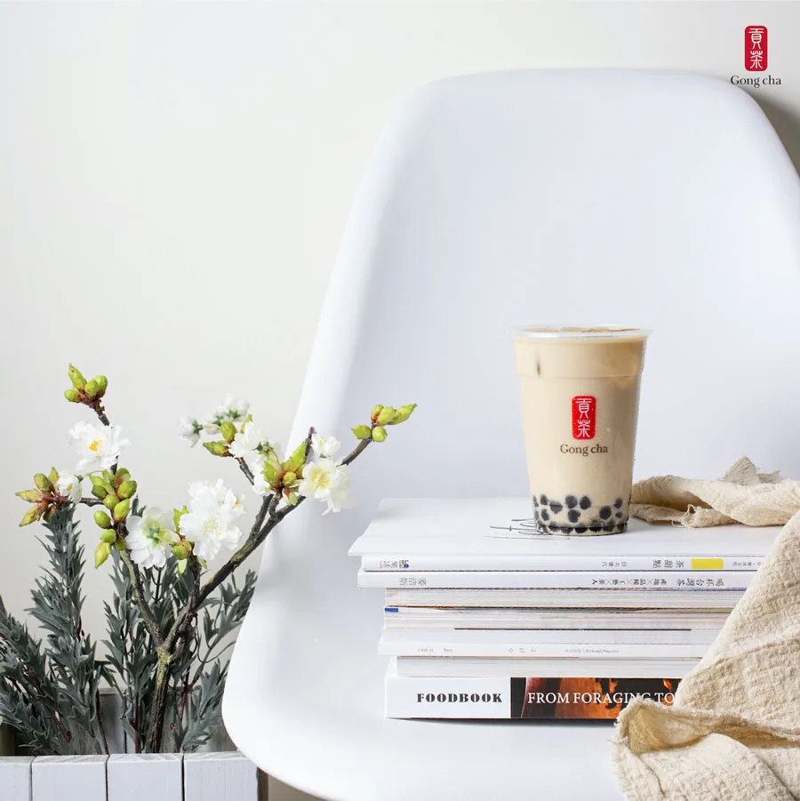 A cup of bubble tea on stacked books beside a flowering branch, all placed on a white chair against a plain background.