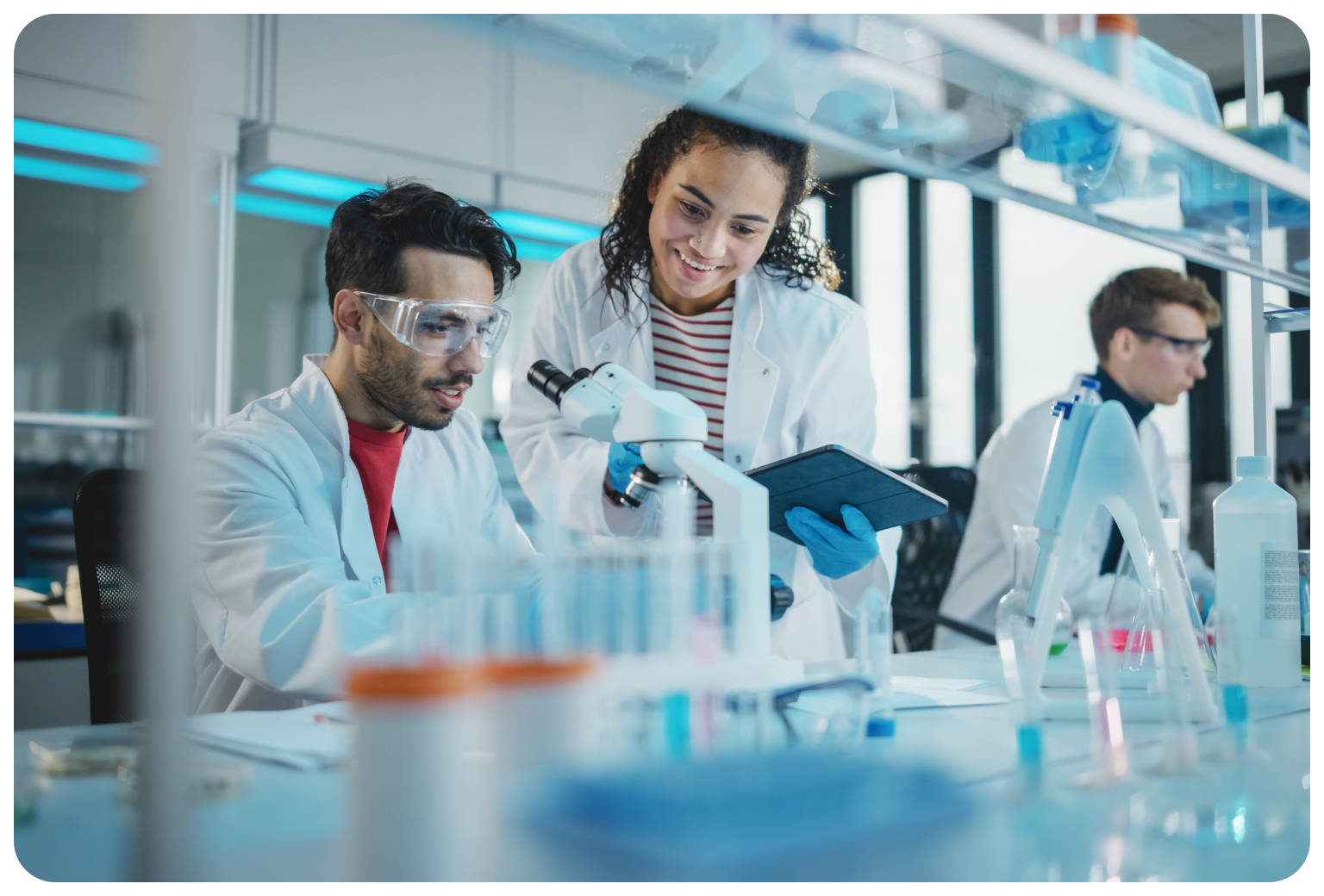 Scientists in lab coats, one using a microscope, another holding a tablet, smiling and discussing findings in a laboratory.