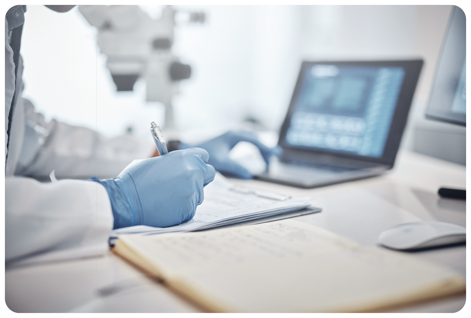 Scientist wearing gloves writes notes in a lab notebook with a laptop and microscope in the background.