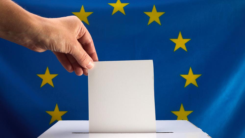 EU flag in background, hand putting slip into ballot box in foreground