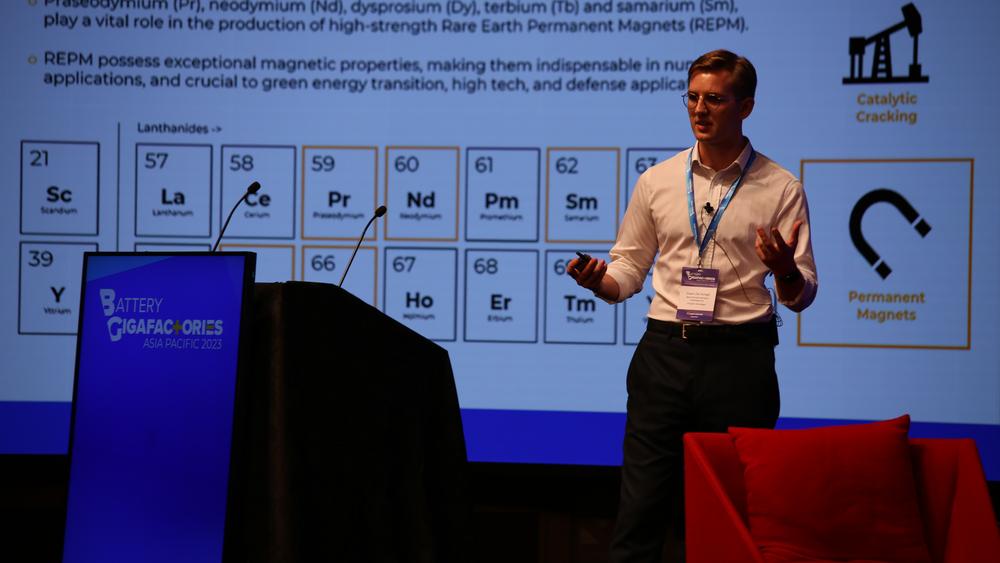 Daan de Jonge speaking on a stage with a presentation slide behind him showing rare earths on the periodic table