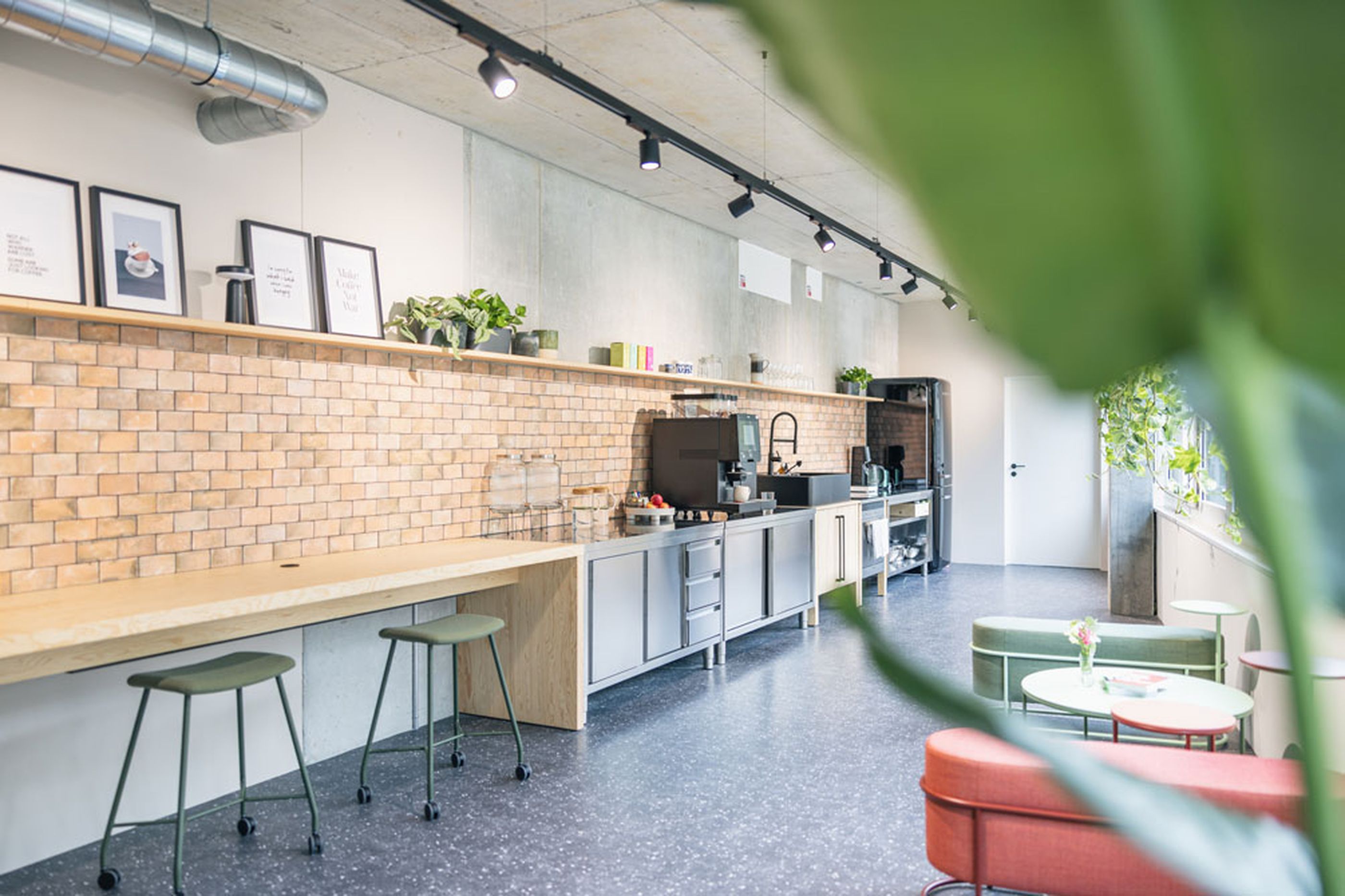 Belebter Bürobereich mit hoher Bar-Arbeitszeile, Backsteinwand und üppigen Pflanzen entlang der Fensterfront.