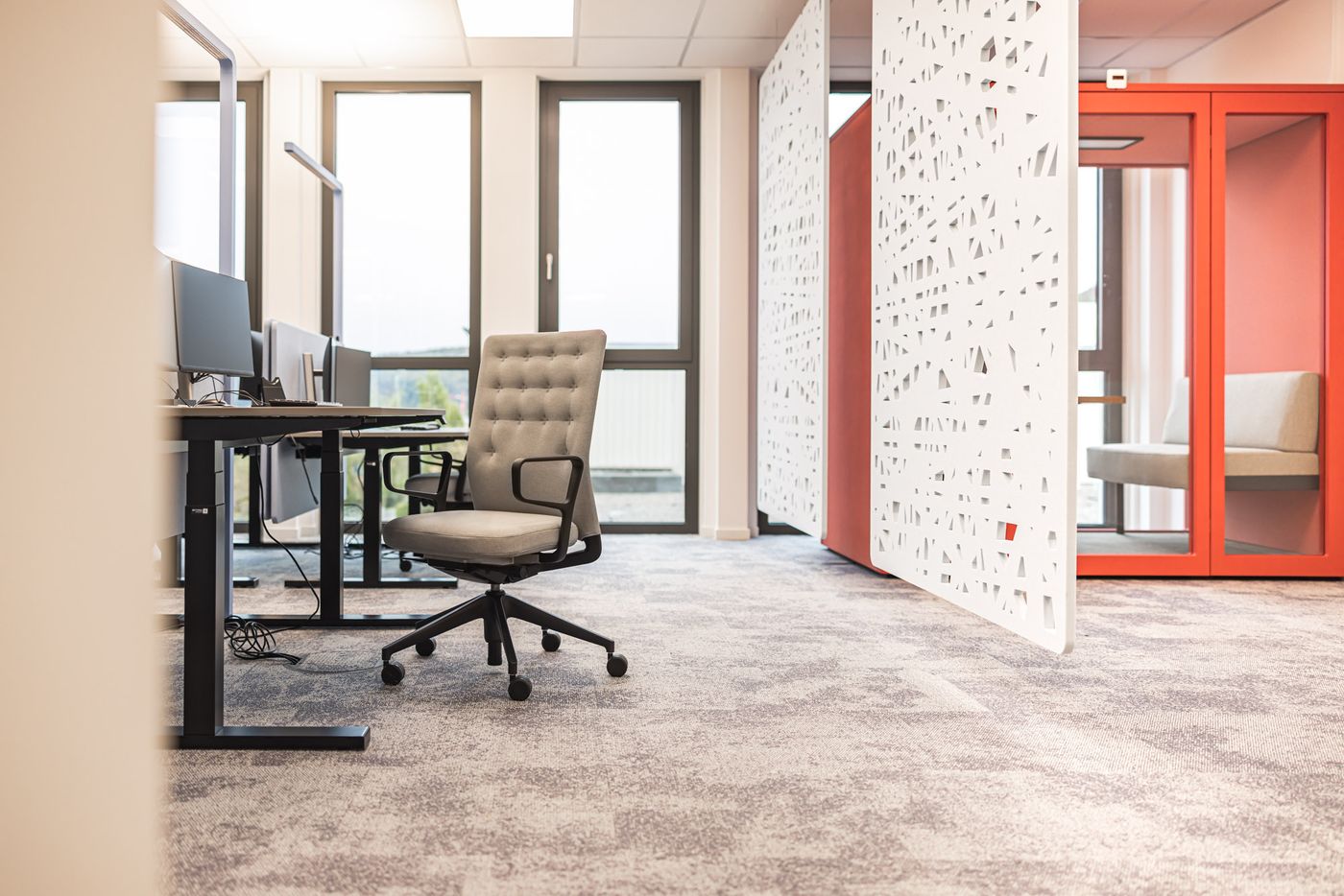 Höhenverstellbare Tische, graue Teppichwelt, weiße Akustiktrenner und auffällige rote Telefonbox.