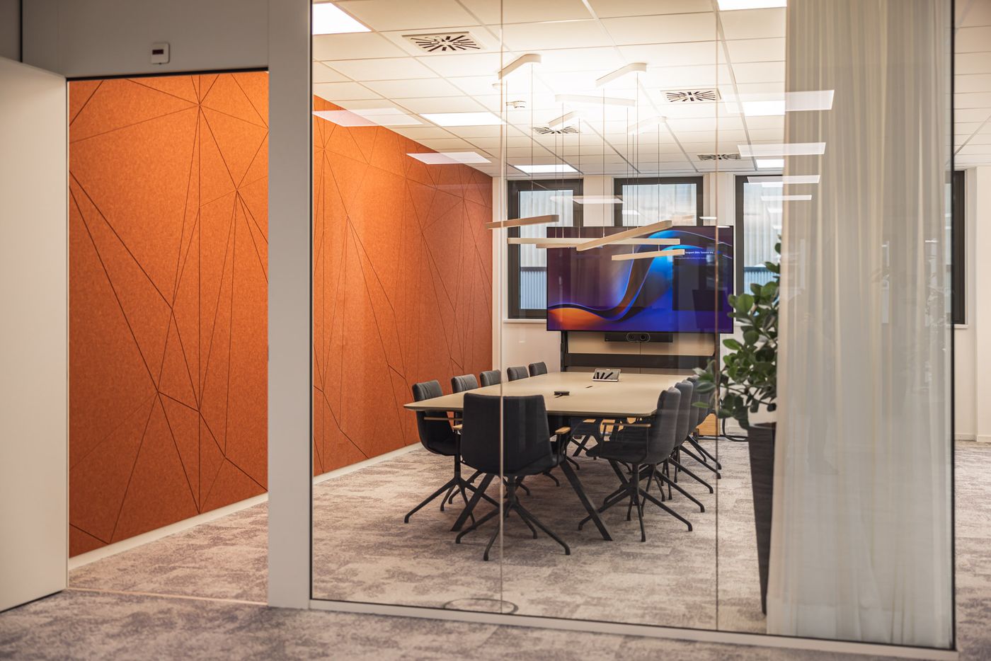 Verglaster Meetingraum mit markanter orangefarbener Akustikwand und großem Screen. Modern, clean, hochwertig.