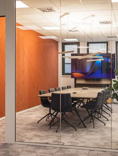 Verglaster Meetingraum mit markanter orangefarbener Akustikwand und großem Screen. Modern, clean, hochwertig.