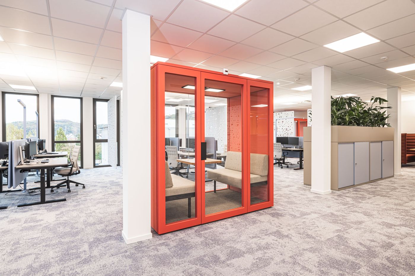 Offene Bürofläche mit auffälliger roter Meetingbox, modernen Arbeitsplätzen und großzügiger Tageslichtbeleuchtung.