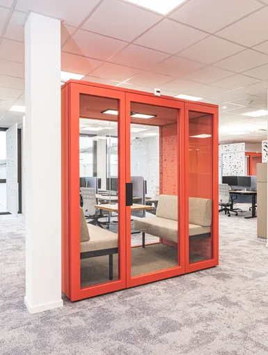 Offene Bürofläche mit auffälliger roter Meetingbox, modernen Arbeitsplätzen und großzügiger Tageslichtbeleuchtung.