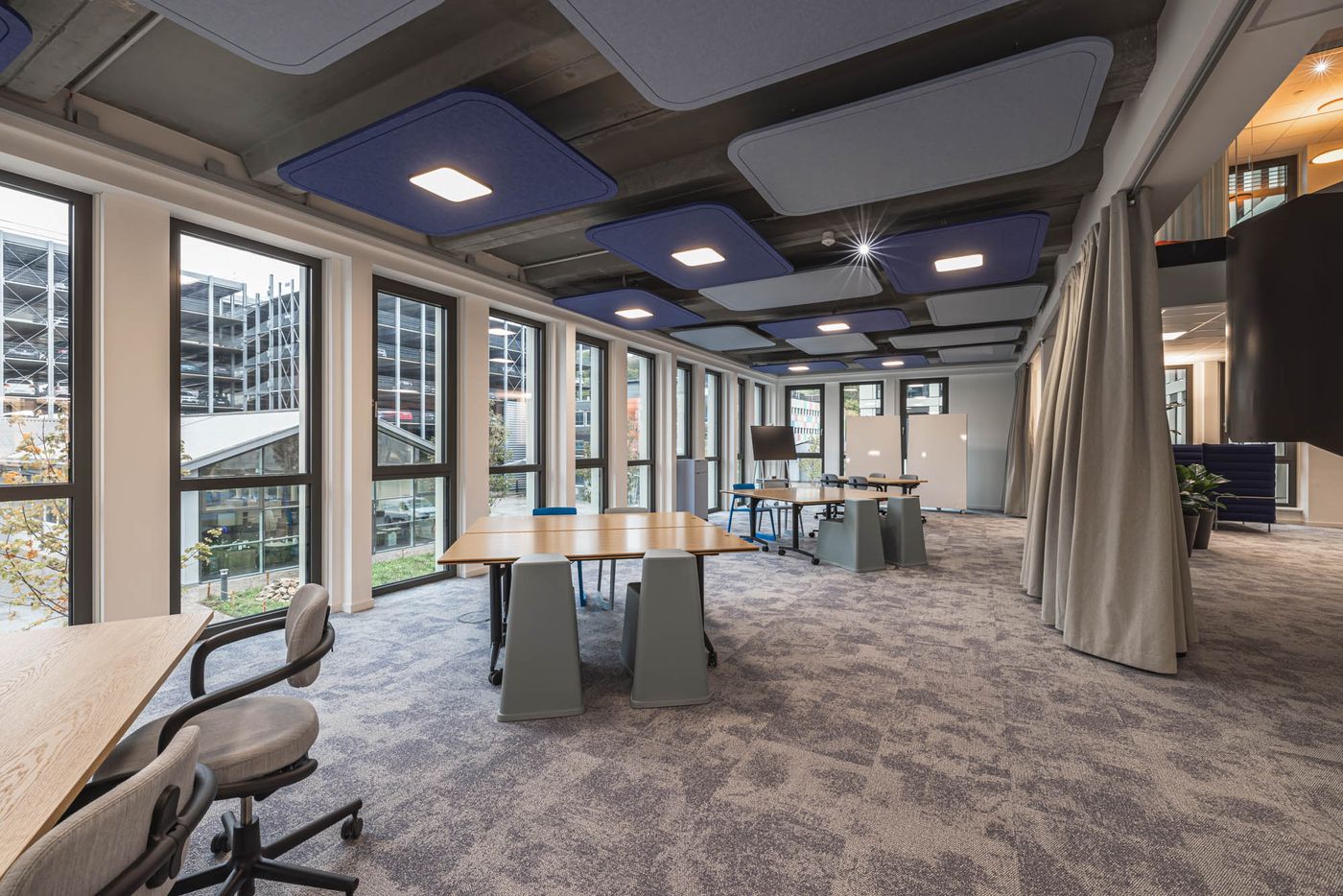 Heller und offener Arbeitsbereich mit großen Fensterfronten, modularen Tischen und akustischen Deckenpaneelen.