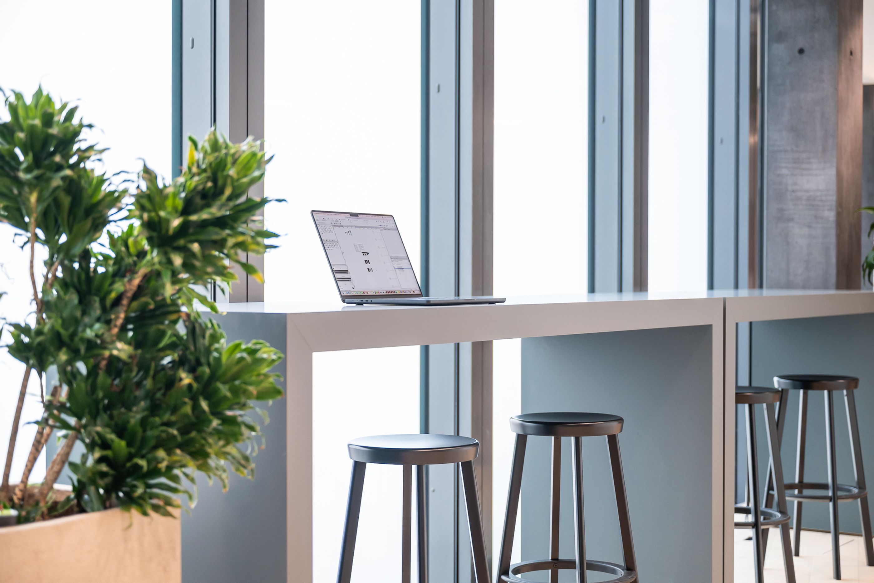 Heller Arbeitsplatz mit hohem Tisch, schwarzen Barhockern und aufgeklapptem Laptop an einer Fensterfront.
