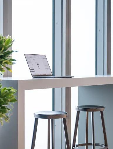 Heller Arbeitsplatz mit hohem Tisch, schwarzen Barhockern und aufgeklapptem Laptop an einer Fensterfront.
