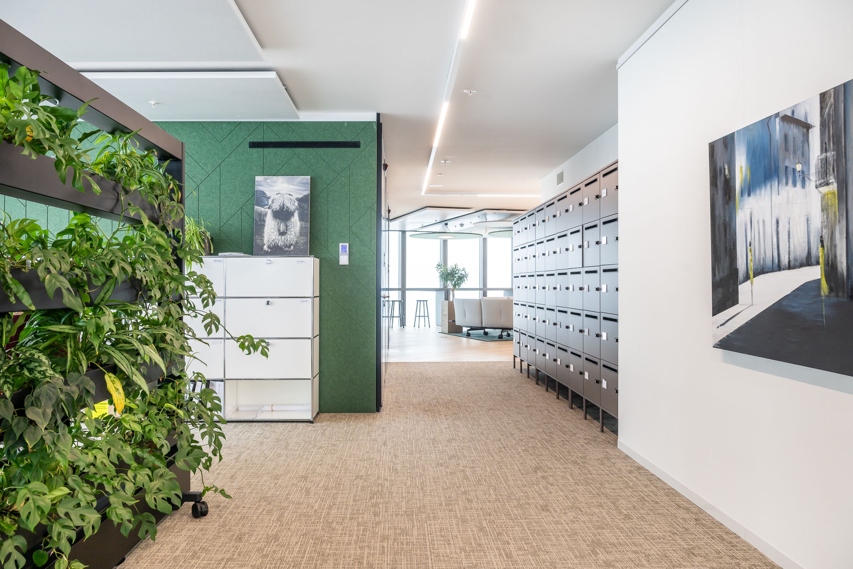 Büroflur mit grüner Akustikwand, mobiler Pflanzenwand, weißen Lockern und großformatigen Wandbildern.