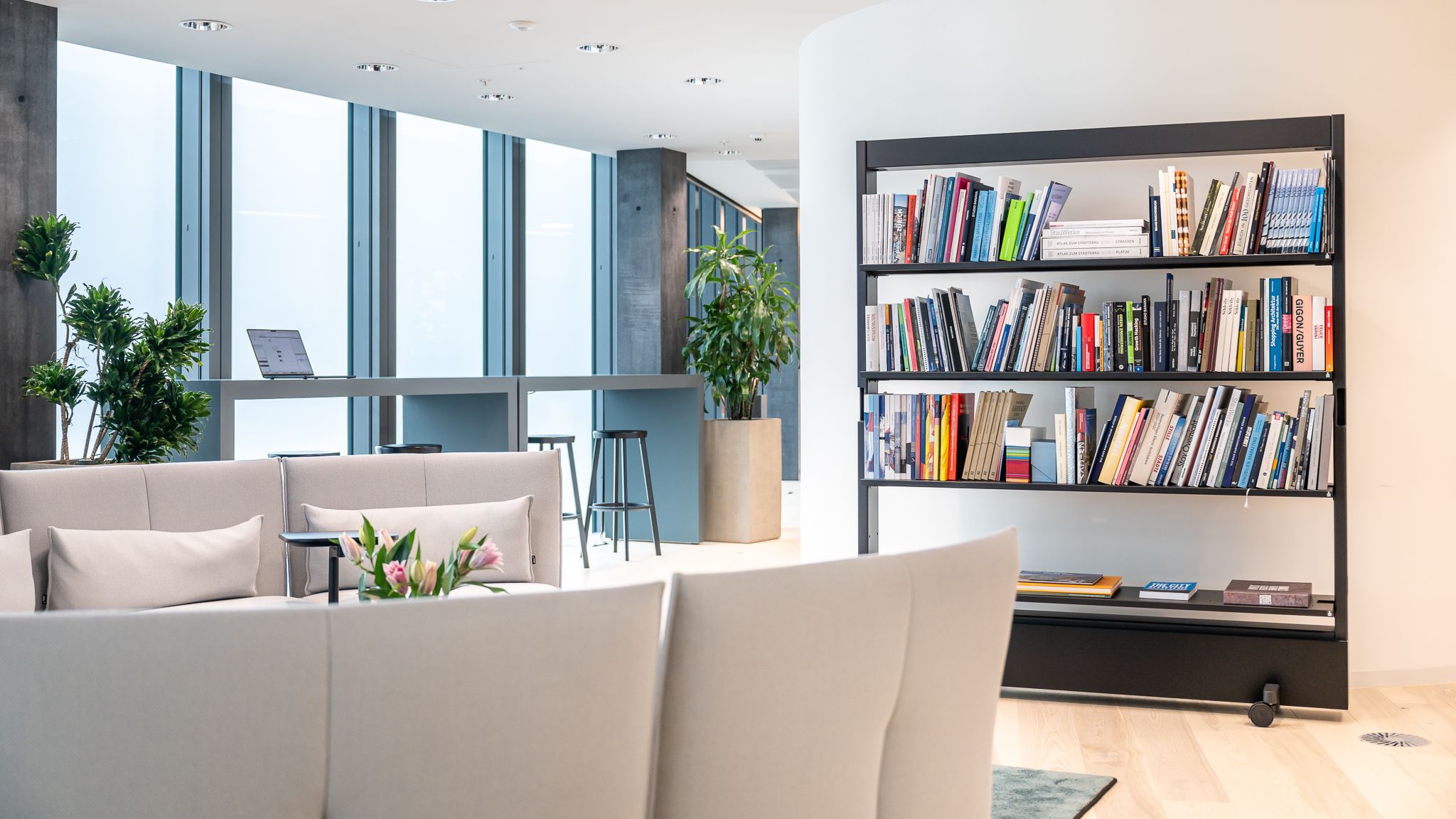 Moderner Loungebereich mit hellen Sofas, großem Bücherregal und hohen Bartischen vor bodentiefen Fenstern.