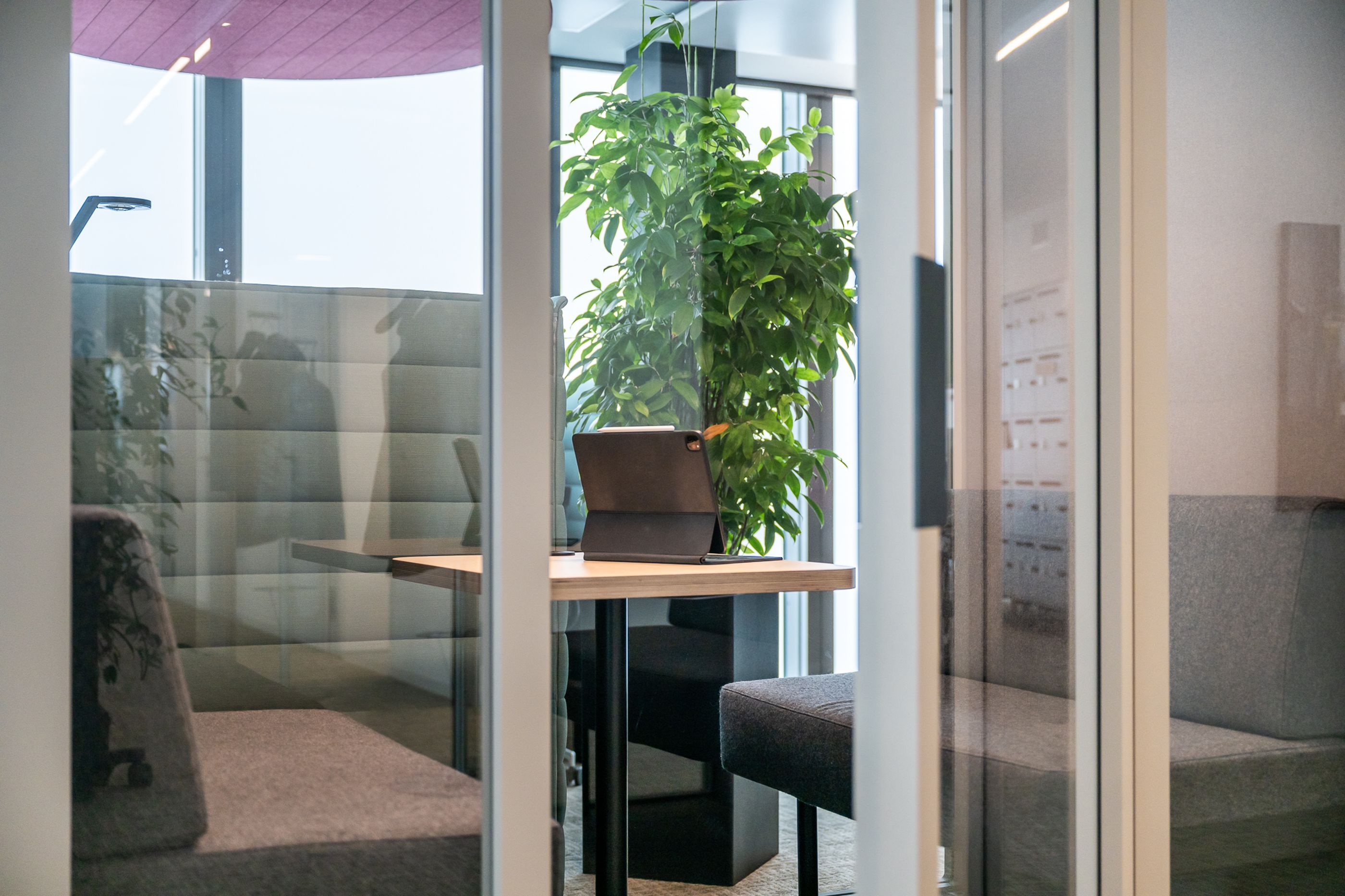 Kleiner Fokusraum mit gepolsterten Booths, Holztisch und Tablet, umgeben von Glaswänden und großer Pflanze.