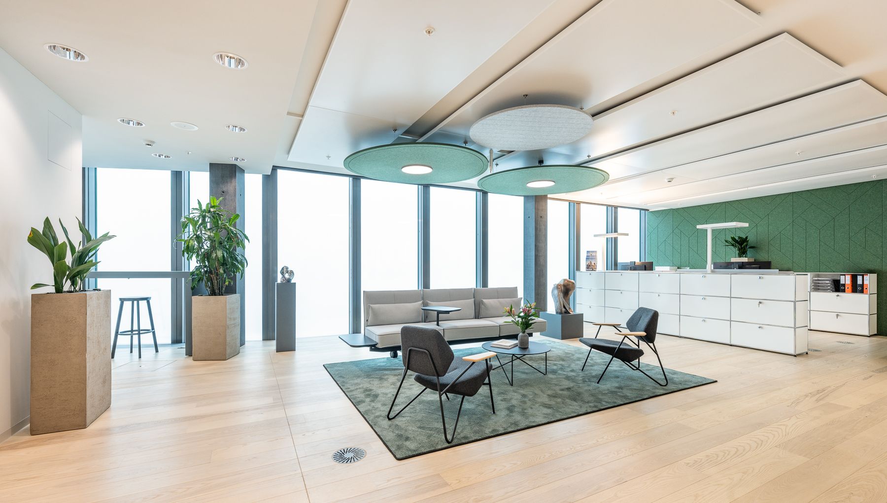 Heller Teamarbeitsbereich mit mehreren Monitoren, grüner Akustikwand und mobiler Pflanzenwand als Raumteiler.