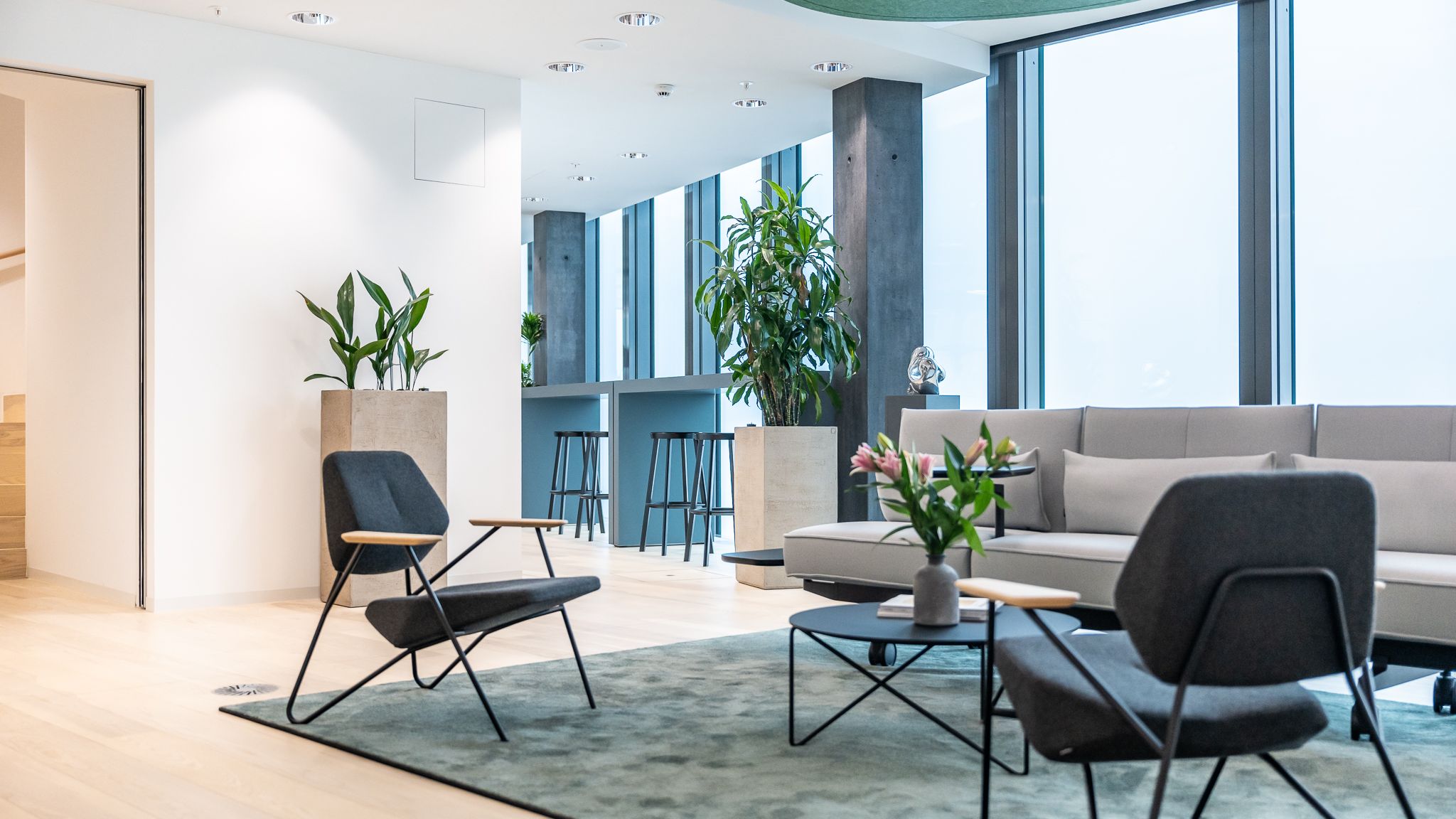 Gemütliche Sitzecke mit Sesseln und Sofa, flankiert von großen Pflanzen und Blick auf hohen Barbereich am Fenster.